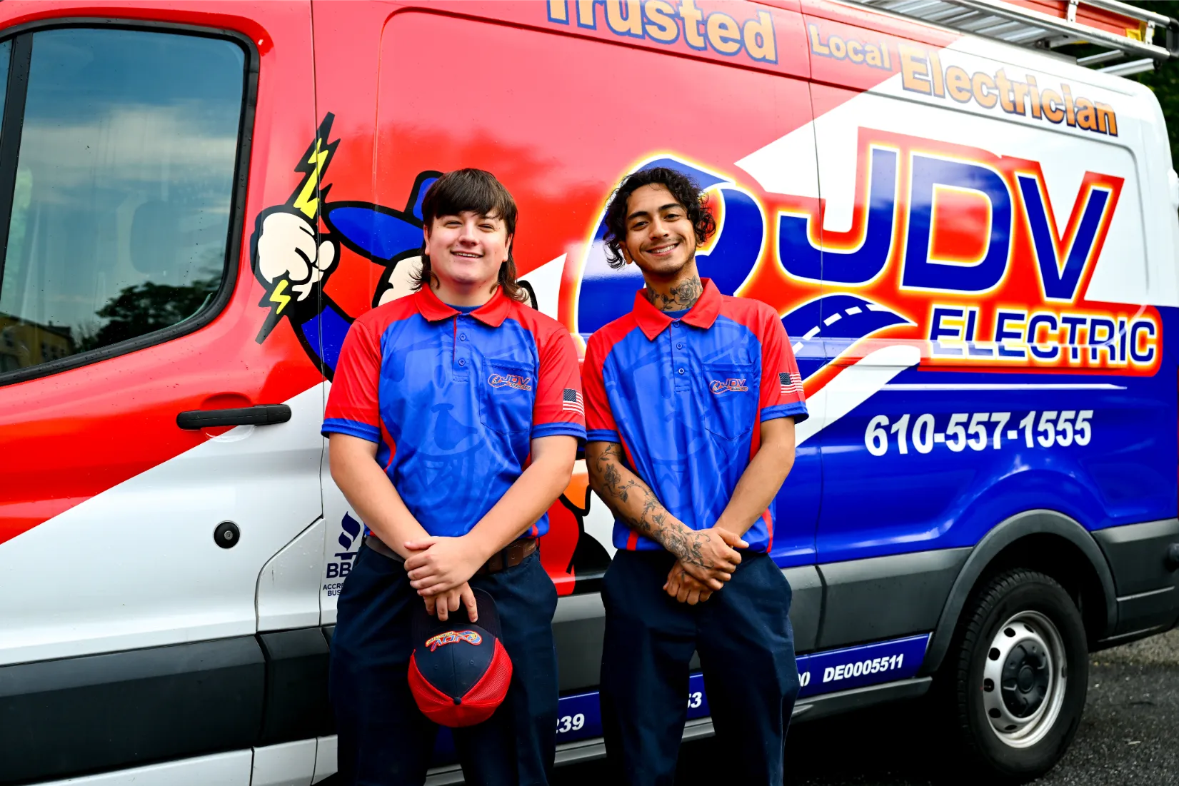 Six JDV Electric employees standing side-by-side together with hands clasped in front of them, standing in a parking lot with JDV service trucks parked behind them.